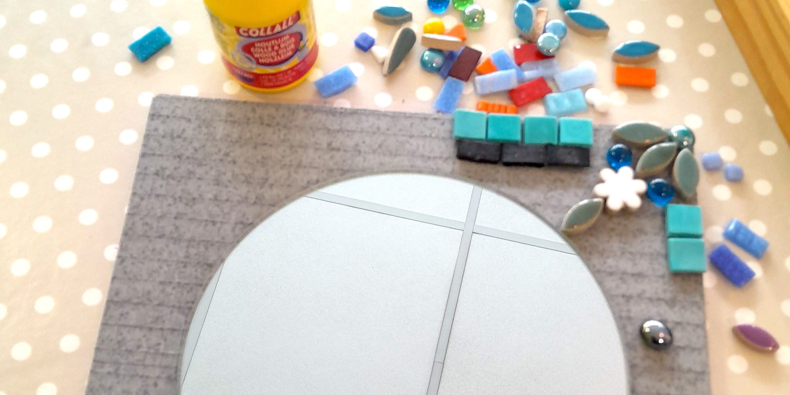 a round mirror sitting on a piece of square tile backer board, with some mosaic tiles and glue around it, waiting to be glued on.