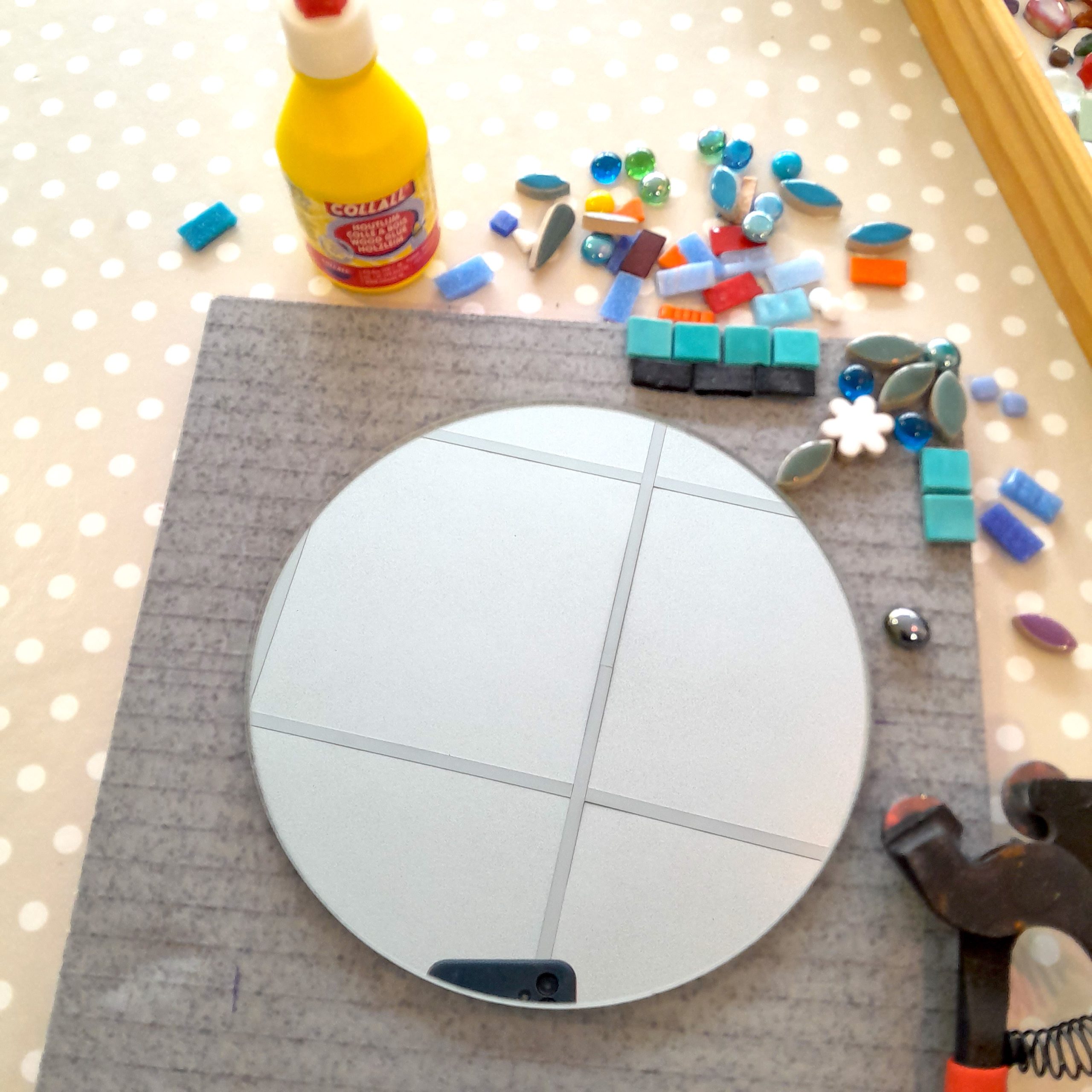 a round mirror sitting on a piece of square tile backer board, with some mosaic tiles and glue around it, waiting to be glued on.