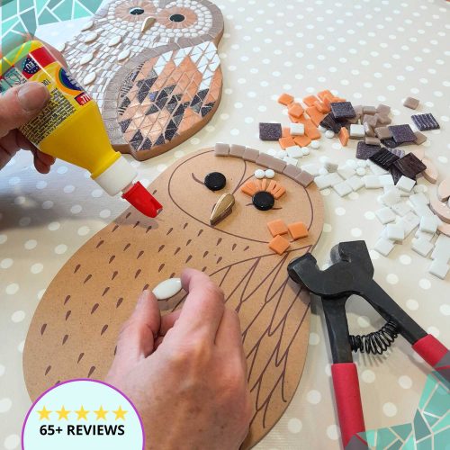 wooden owl shaped MDF board on a table with glue, mosaic tile cutters and mosaic tiles surrounding it, as though it is a mosaic work in progress. A finished example of the large mosaic owl kit is in the background.